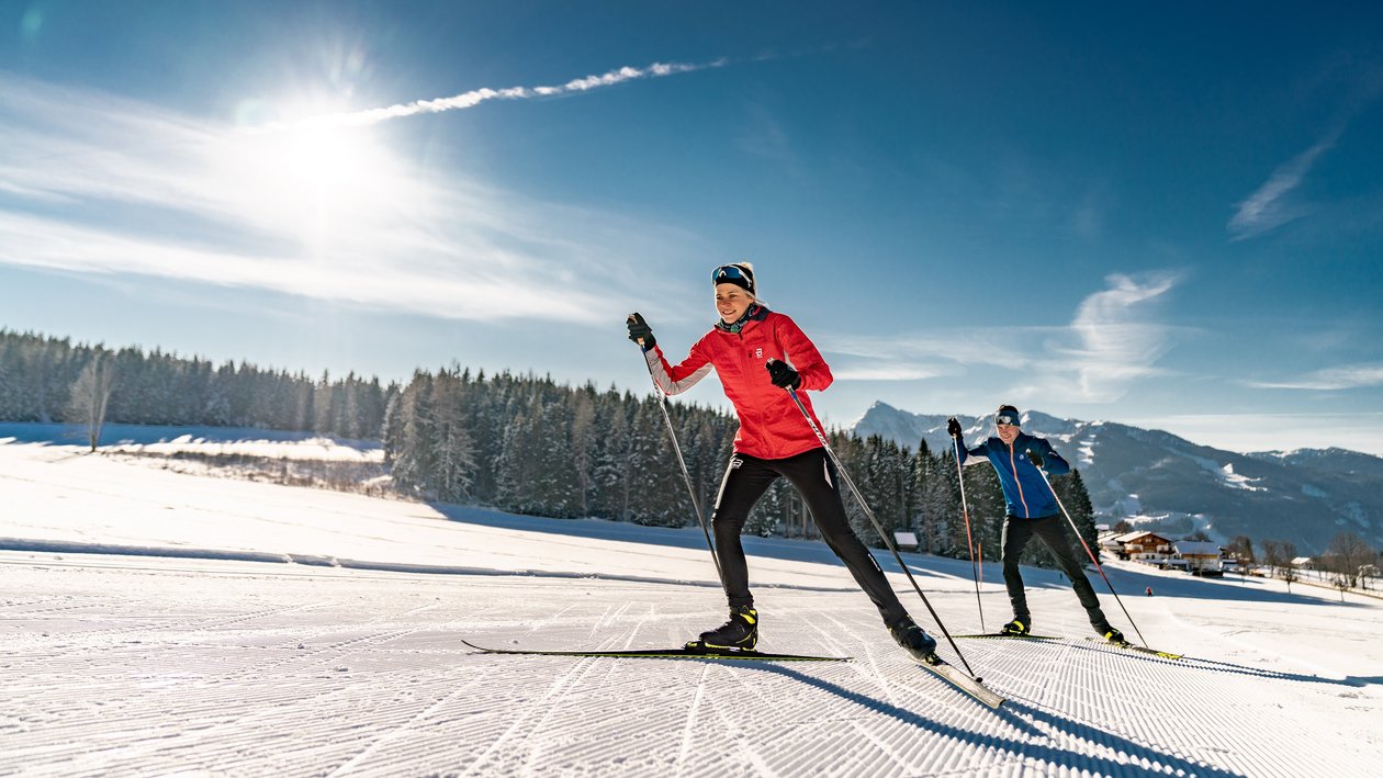 Langlaufen im Winterparadies Zwei Personen beim Langlaufen auf einem verschneiten Weg unter blauem Himmel und Bergen
