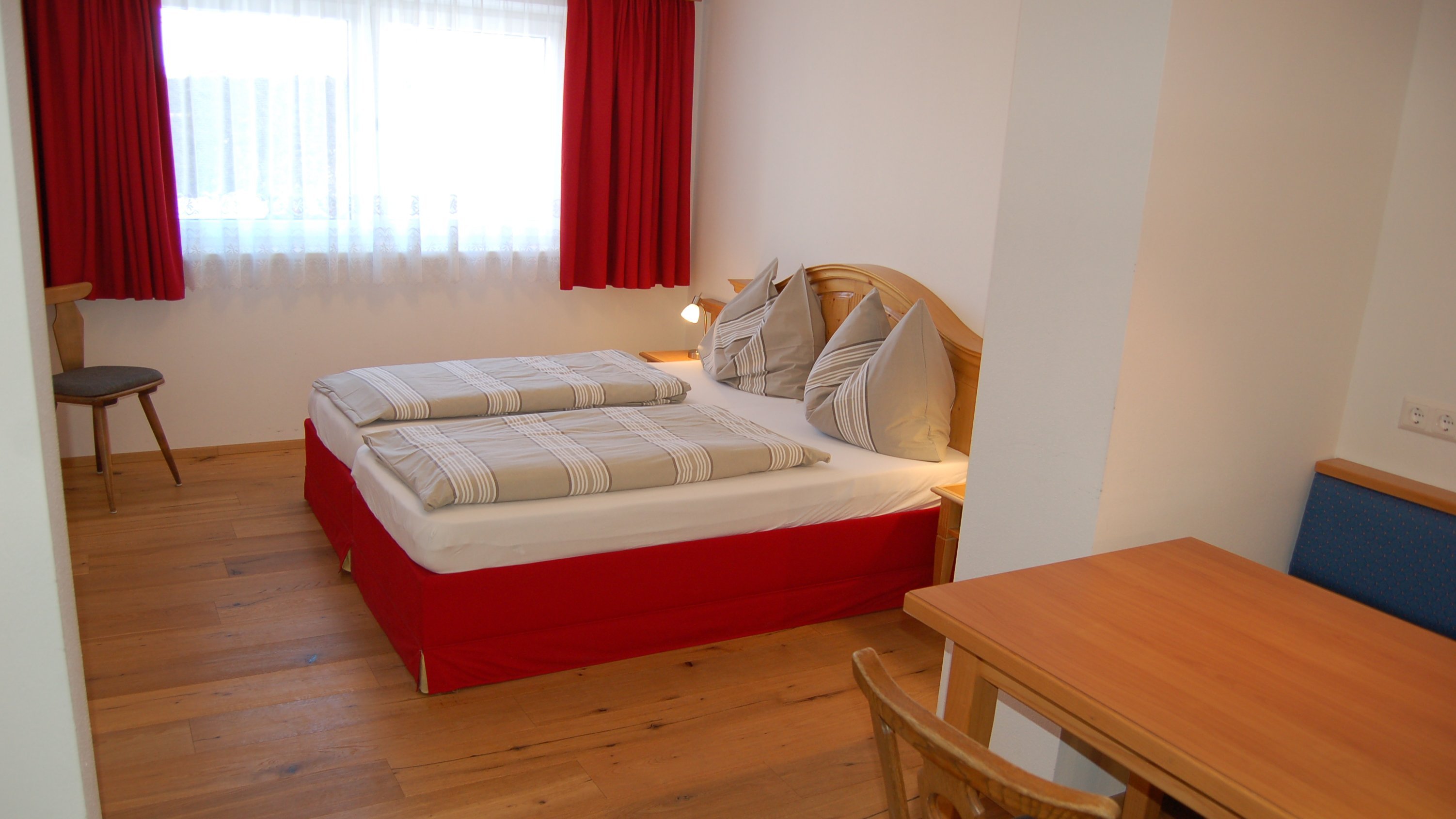 Hotelzimmer mit zwei Betten, Holztisch und Fenster mit roten Vorhängen in einem modernen Design.
