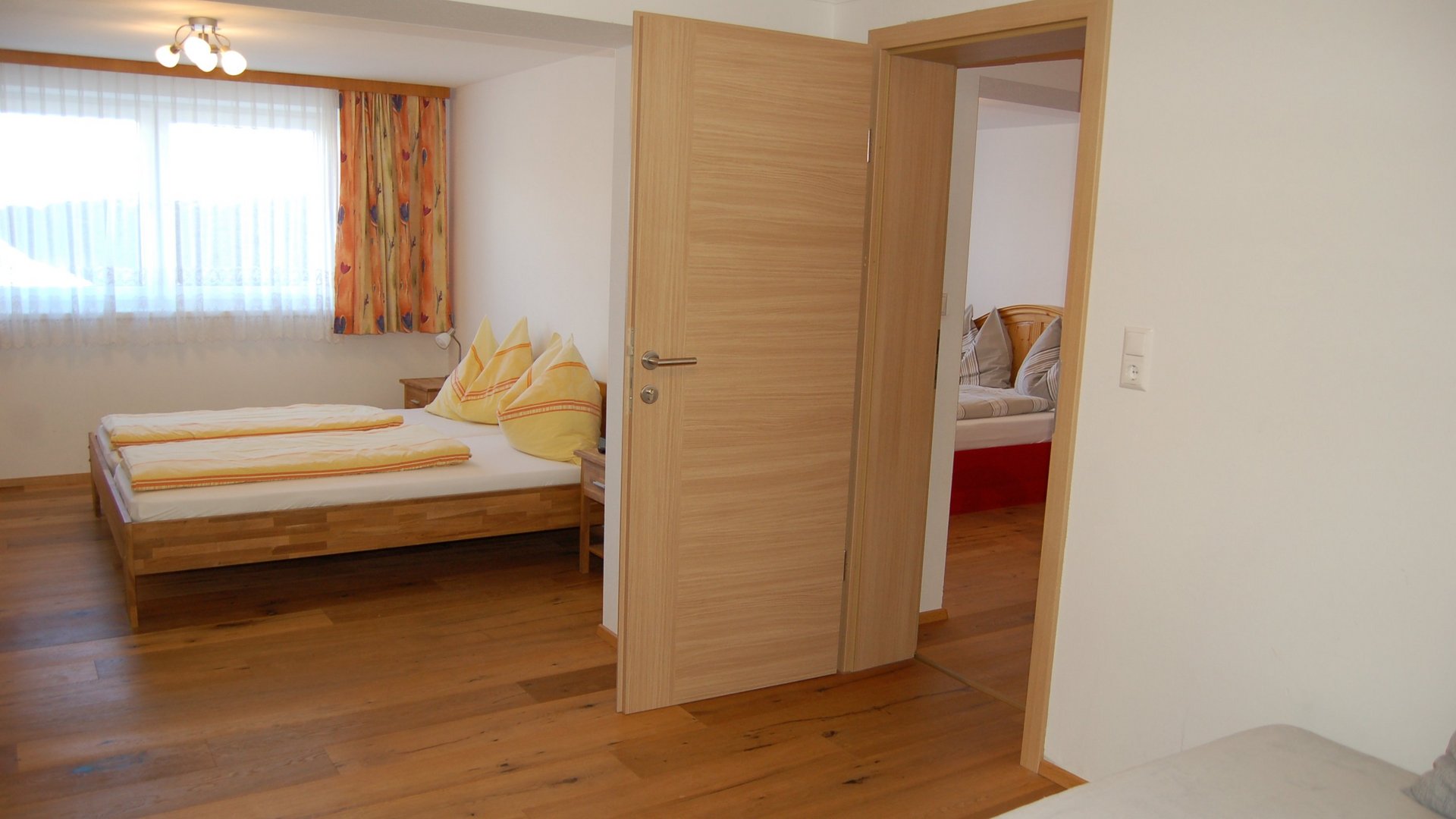 Helle Hotelzimmer mit zwei Doppelbetten und Holzboden, Durchgang zu einem weiteren Raum sichtbar
