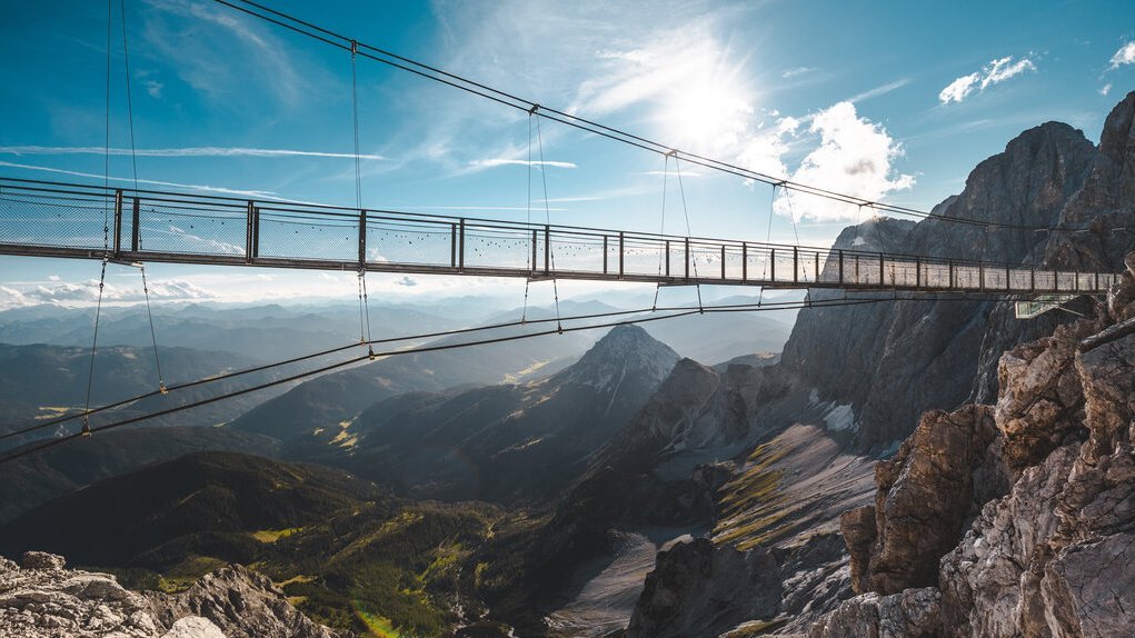Eine schwebende Brücke über eine Berglandschaft mit klarem Himmel und Sonnenlicht.