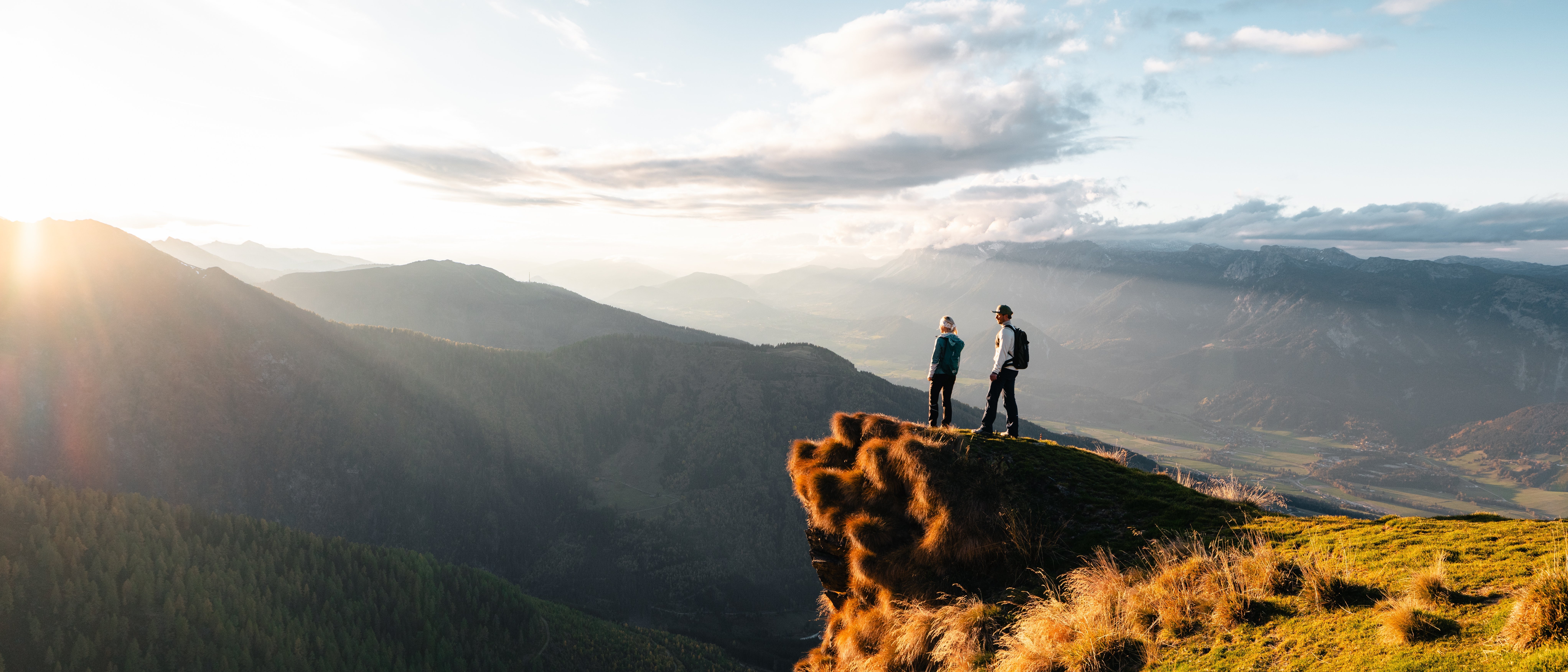 Zwei Wanderer stehen auf einem felsigen Aussichtspunkt und blicken auf eine Berglandschaft bei Sonnenuntergang.