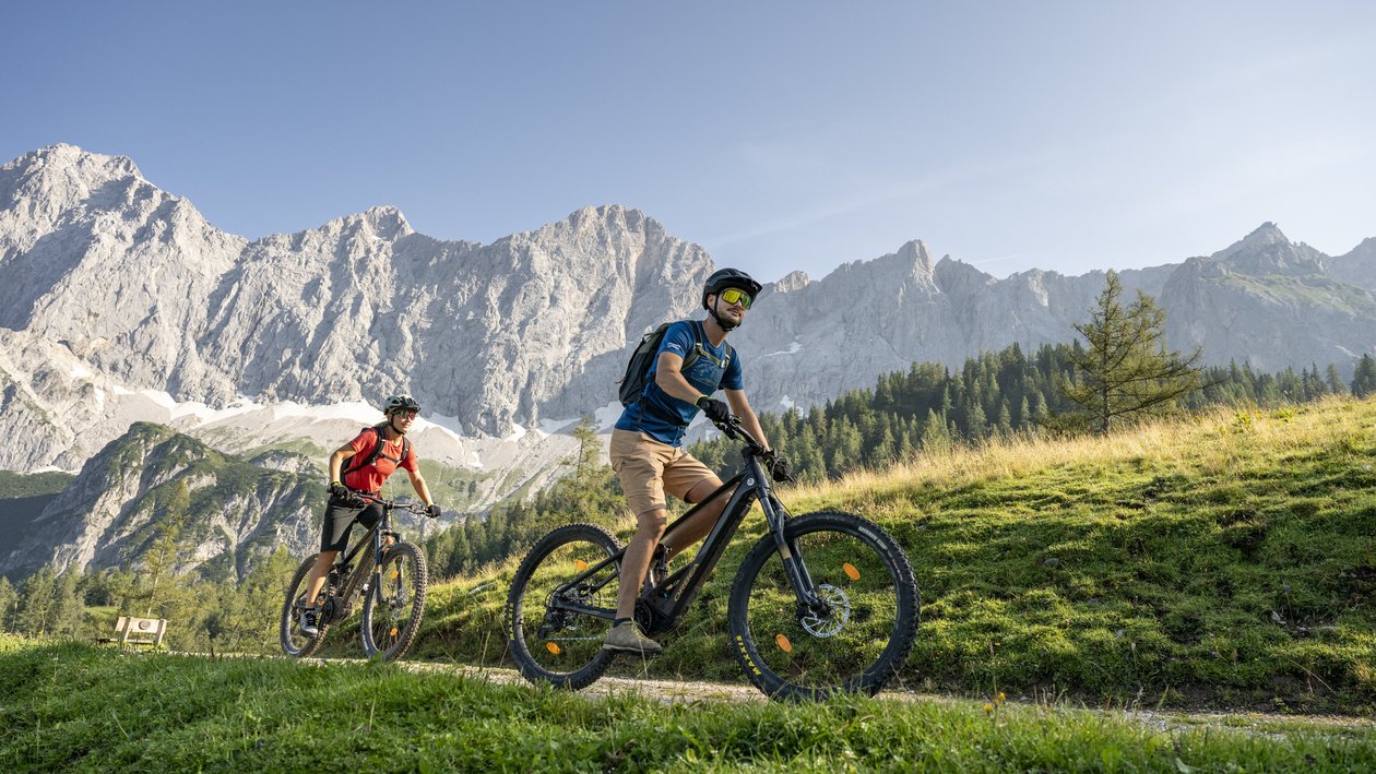 Zwei Personen fahren mit Mountainbikes auf einem schmalen Trail zwischen grünen Bergen.