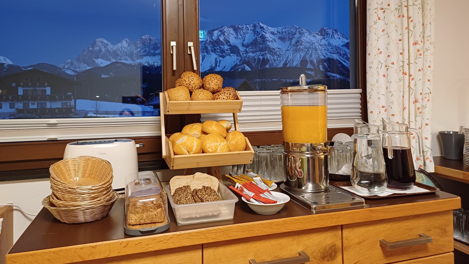 Buffet mit verschiedenen Backwaren und Getränken vor einer Bergkulisse. 1. Verschiedene Brötchen, Croissants und Säfte.