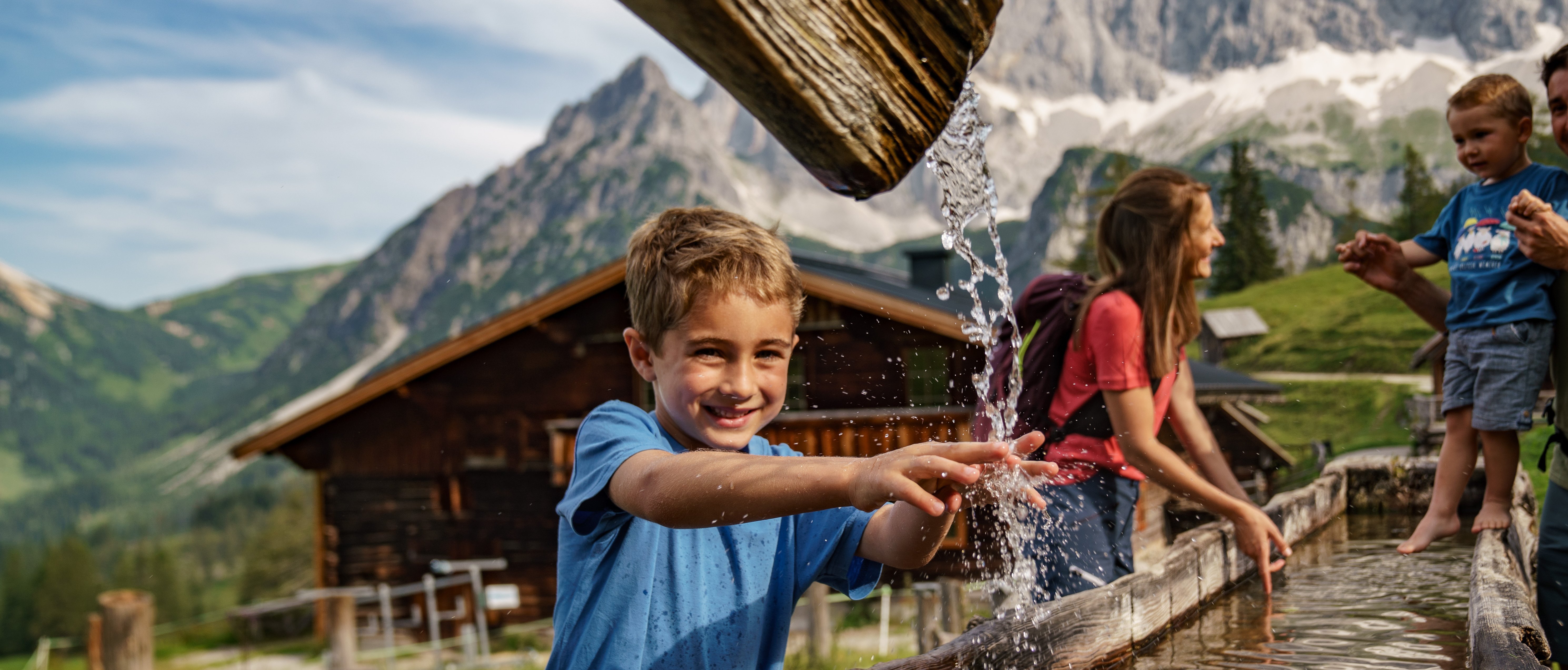 Eine Familie spielt im Wasserbrunnen vor einer bergigen Kulisse mit Kindern und Natur.