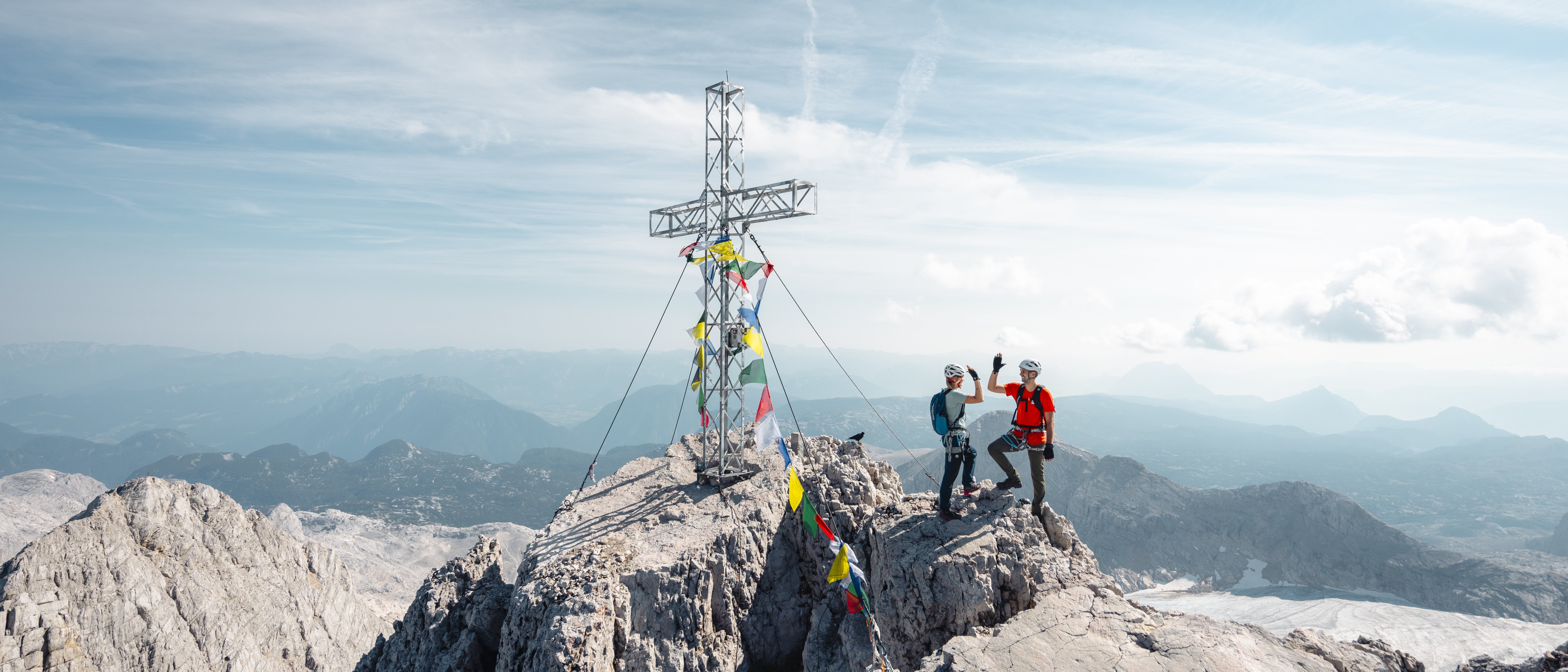 Zwei Bergsteiger stehen an einem Gipfelkreuz, umgeben von bunten Gebetsfahnen mit Panoramablick auf die Landschaft.