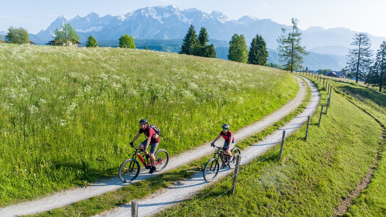Zwei Radfahrer fahren einen gewundenen Pfad in einer grünen Landschaft mit Bergen im Hintergrund.