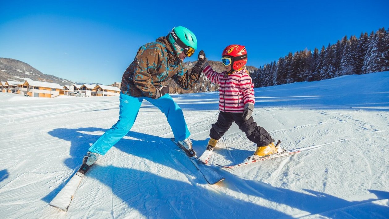 Eine Person und ein Kind fahren Ski auf einer verschneiten Piste und interagieren spielerisch.