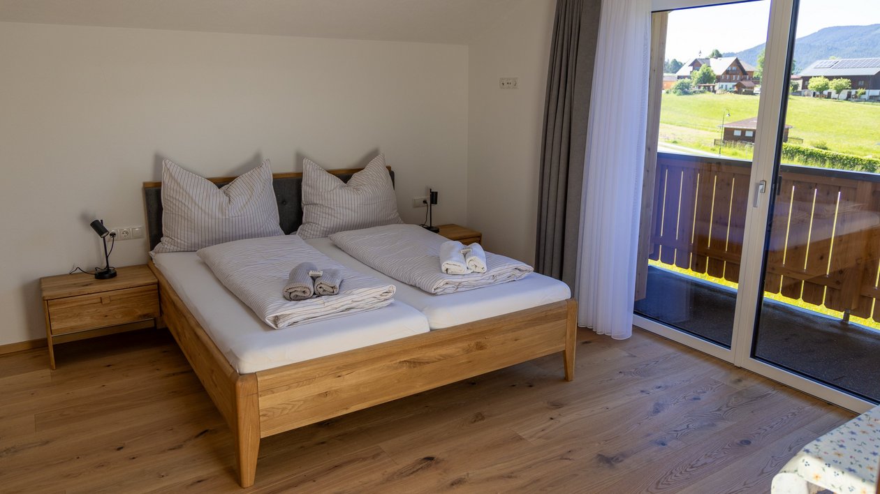 Bright hotel bedroom featuring a double bed, wooden furniture, and balcony access to an outdoor view