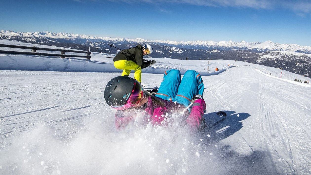 Zwei Personen beim Skifahren auf einer schneebedeckten Piste vor Berglandschaft und blauem Himmel.