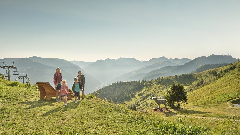 Familie auf einer Wiese mit Blick auf Berge und Skilifte im Hintergrund, Holzbank sichtbar