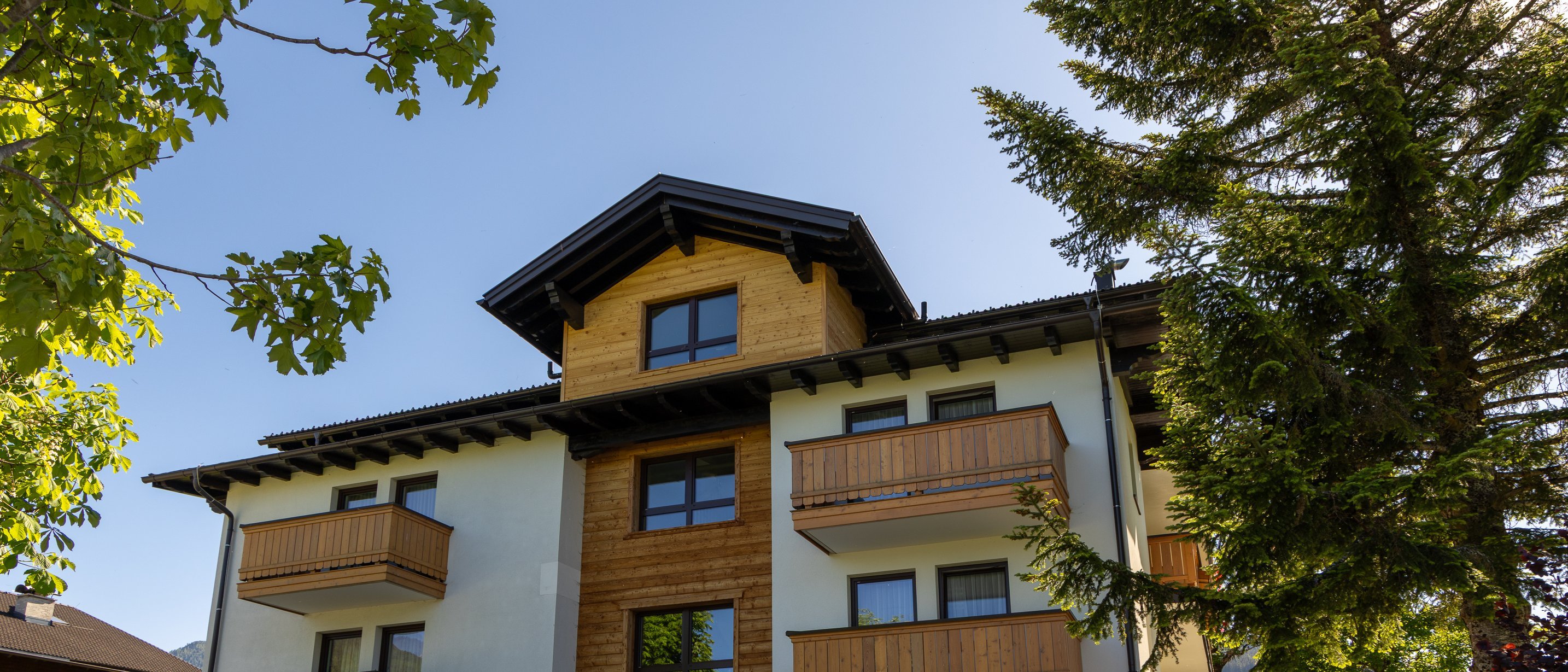 Dreistöckiges Wohngebäude mit Holz- und Weißverkleidung, Balkonen und umgeben von Grünflächen.