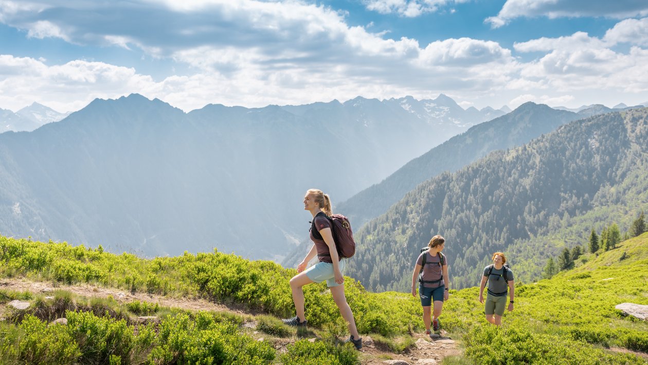 Drei Wanderer auf einem Pfad in einer bergigen Landschaft mit üppigem Grün und fernen Gipfeln unter teils bewölktem Himmel.