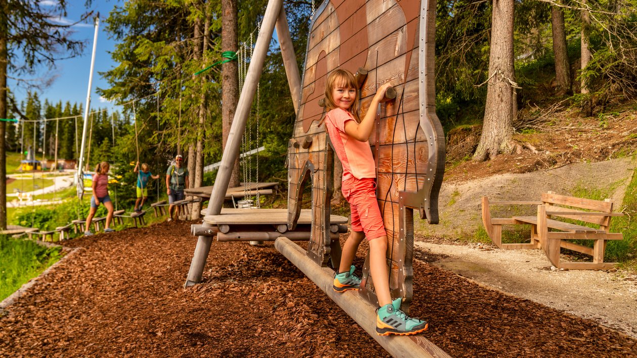 Kinder spielen im Freien auf einem Holzspielplatz mit Kletterstruktur umgeben von Bäumen.