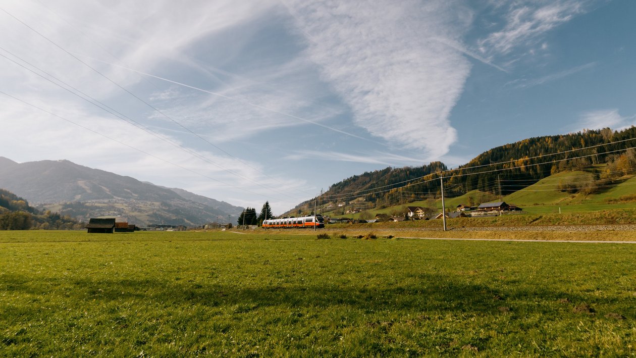 Ein Zug fährt durch ein grünes Feld mit Bergen im Hintergrund und klarem blauen Himmel.