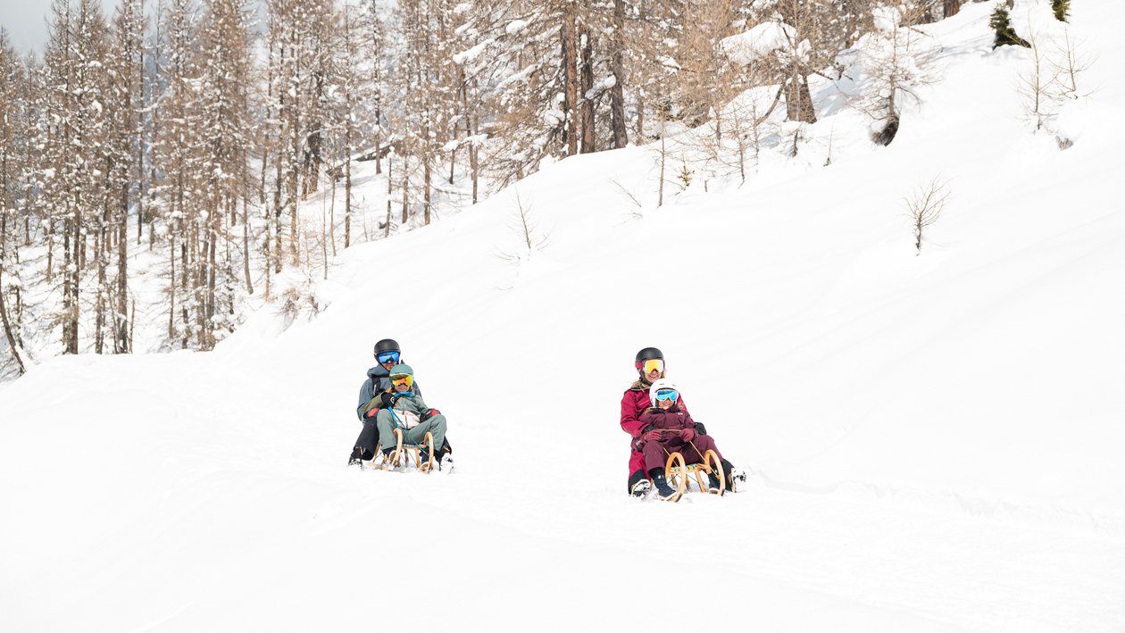 Drei Personen rodeln einen verschneiten Weg entlang, umgeben von Bäumen in einer Winterlandschaft.