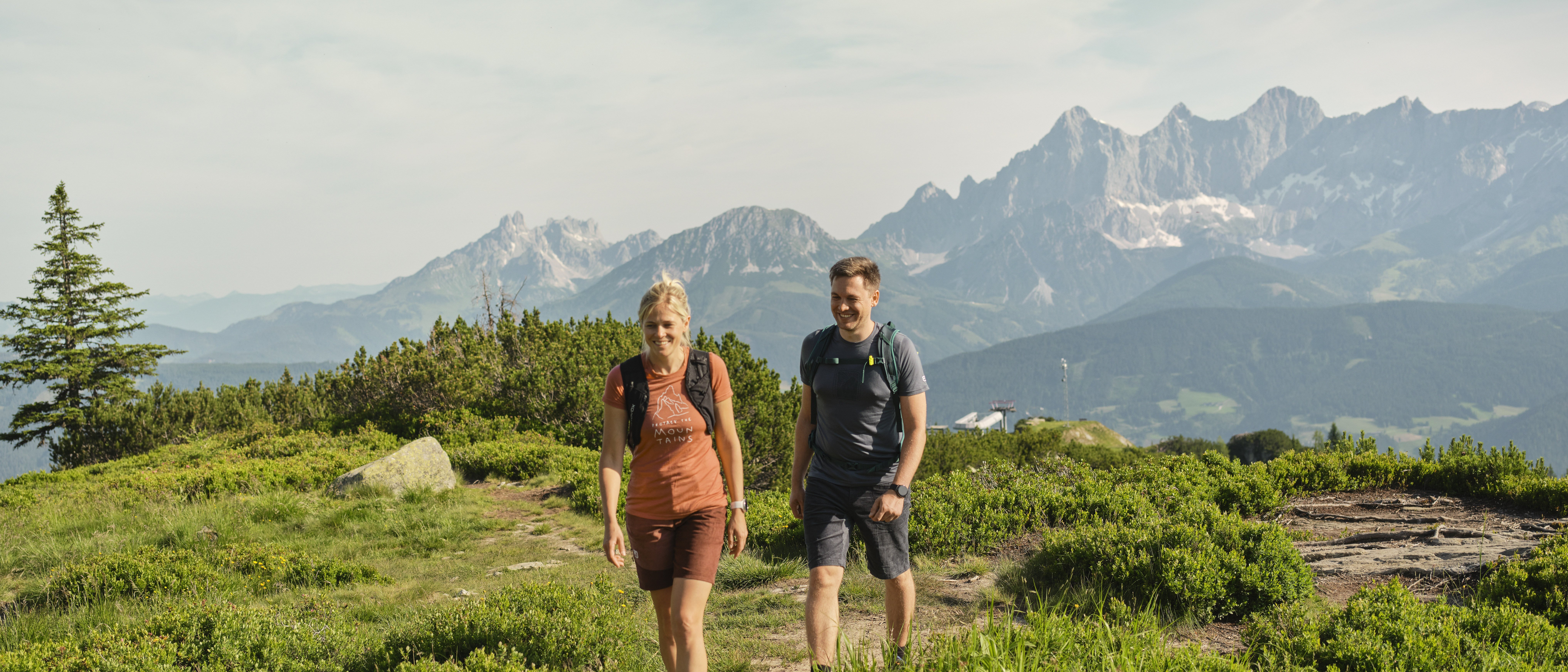 Ein Paar wandert auf einem Pfad umgeben von grünen Pflanzen und Bergen im Hintergrund.