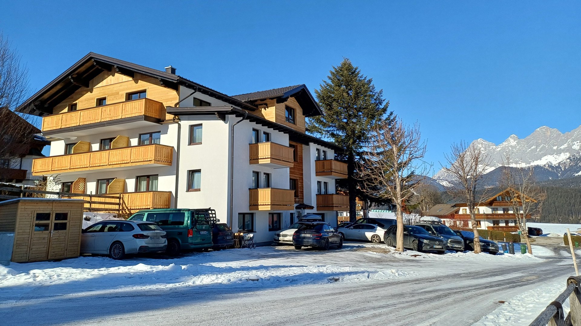 Berghütte umgeben von Schnee mit geparkten Autos und Bergen im Hintergrund unter klarem Himmel